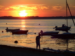 Wasserspiegelung vom Sonnenuntergang in der Eckernförder Bucht (bei Noer), im Vordergrund ein Mensch und mehrer geankerte kleine Boote